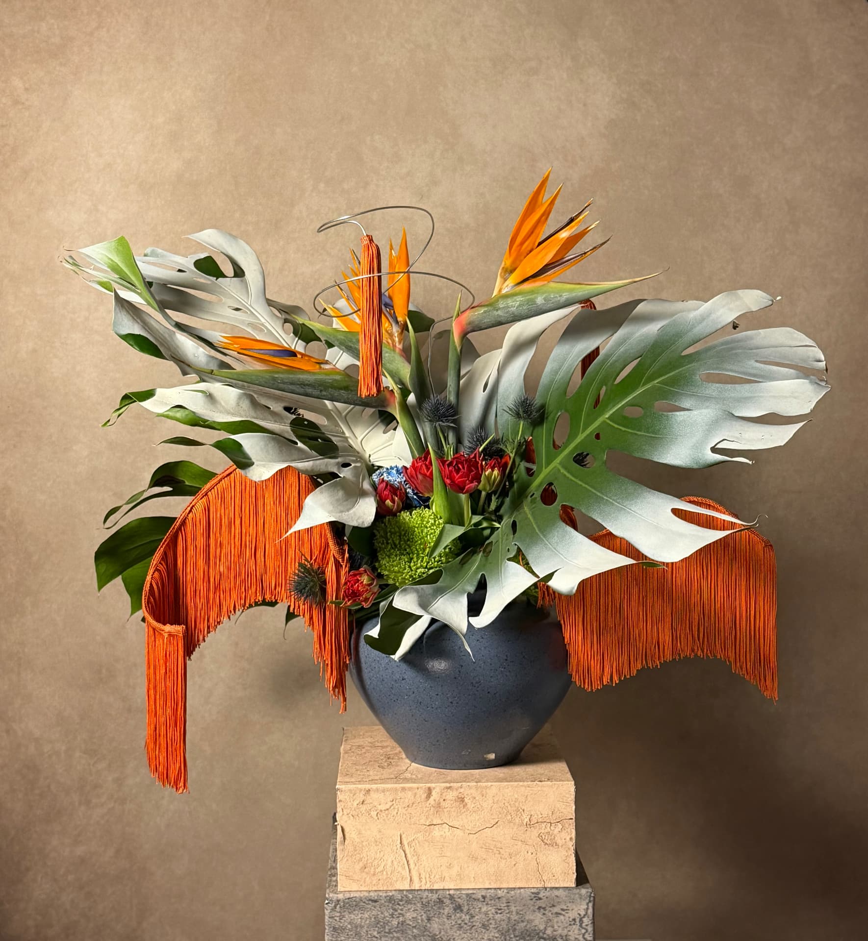 Sculptural arrangement of silvered monstera leaves, bird of paradise, orange fringe and red roses in a slate-blue vessel on a stone plinth